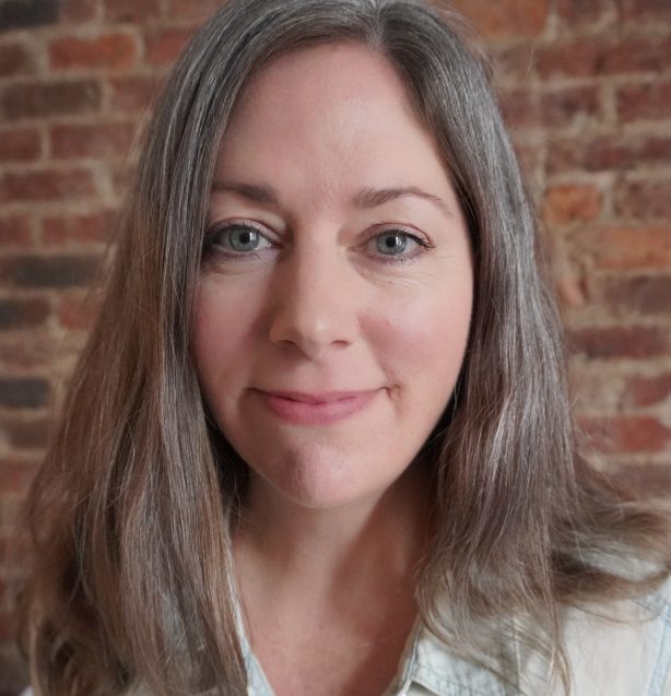Jenny Haufek Headshot - Jenny has long brown hair with sprinkles of silver and wears a pale green shirt, smiling at the camera