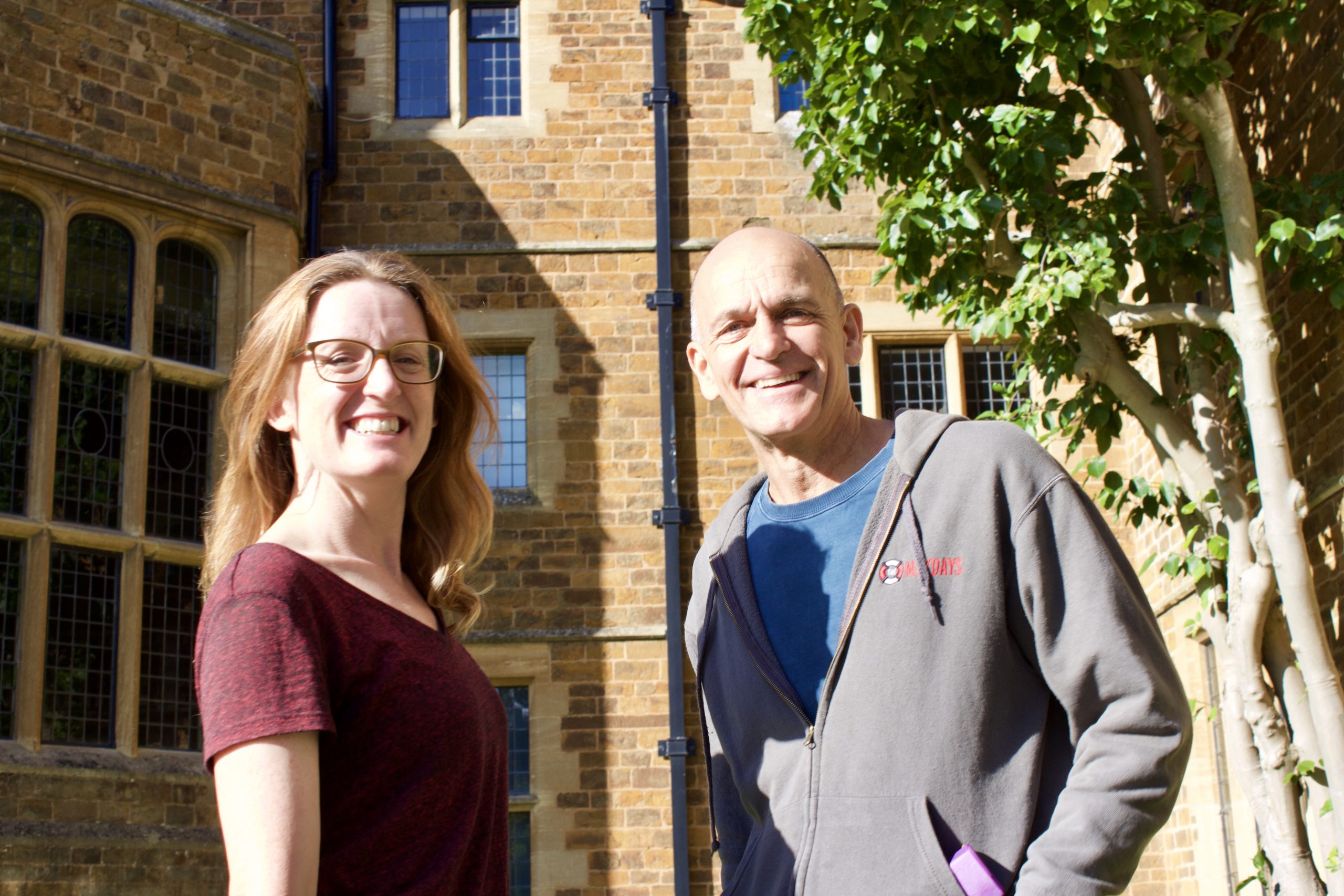 A woman (The Maydays Liz Peters) wears glasses and a big smile with long light brown hair down and a red v-neck t-shirt, besides her stands a man (The Maydays John Cremer) wearing a big smile and a blue t-shirt with a grey maydyas hoodie. Both stand outside a grand building (The Maydays improv retreat venue, Ashorne Hill)